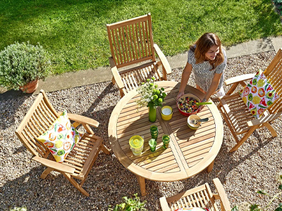 Frau deckt runden Holztisch mit bunten Kissen, umgeben von Grün und Kies
